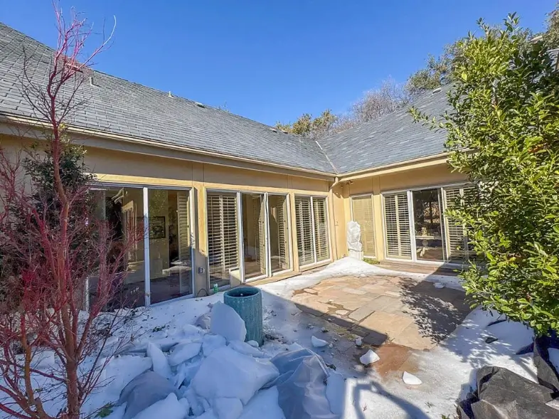 A sunny courtyard with snow patches, glass doors, a blue planter, and surrounding trees.