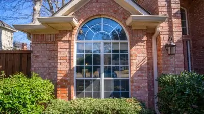 Large arched window with grid panes on a red brick house, bordered by green bushes and outdoor light fixture.